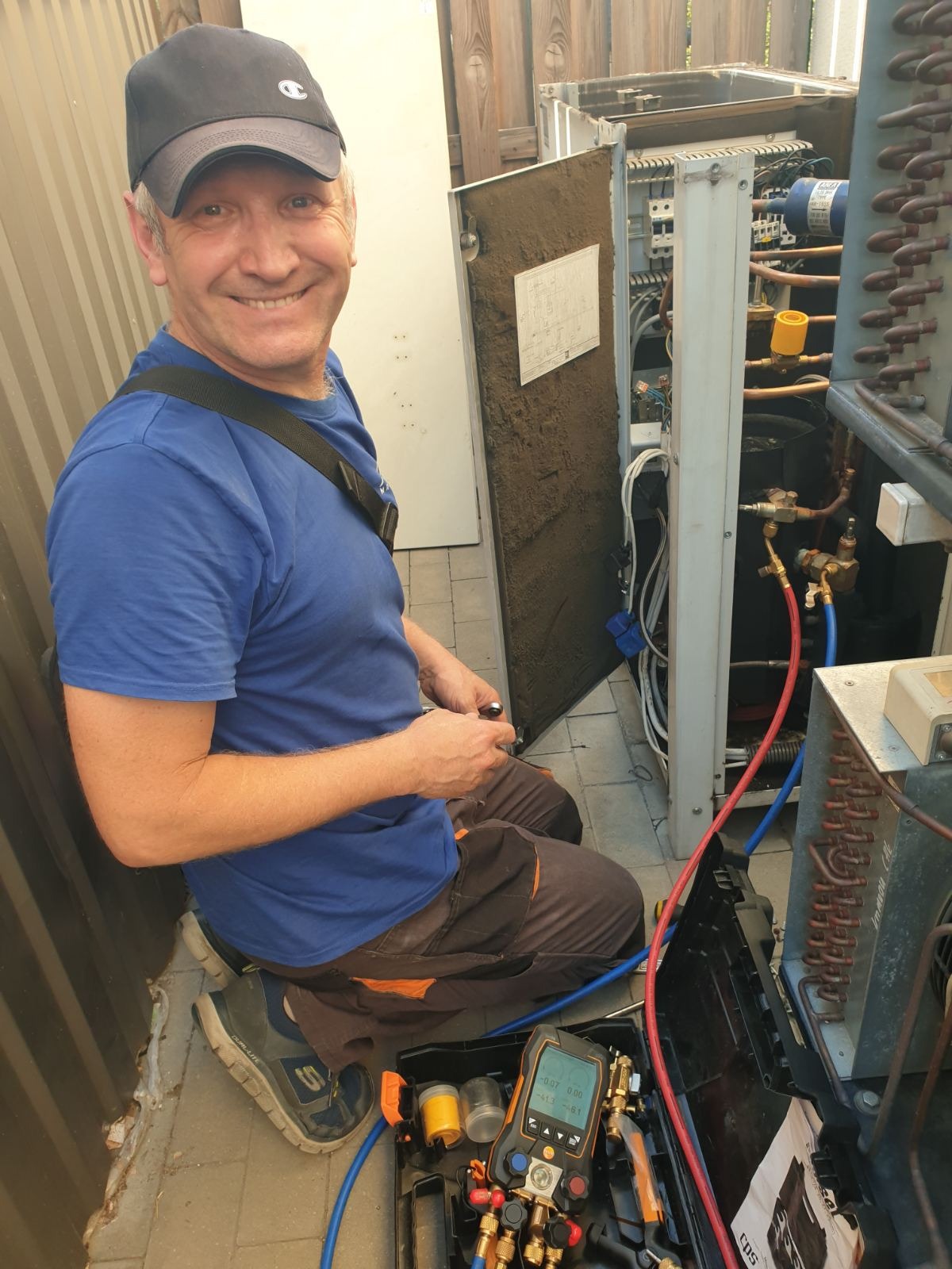 Uśmiechnięty serwisant w czapce naprawia klimatyzator na zewnątrz, używając specjalistycznych narzędzi. Widoczne miedziane rurki i miernik ciśnienia.