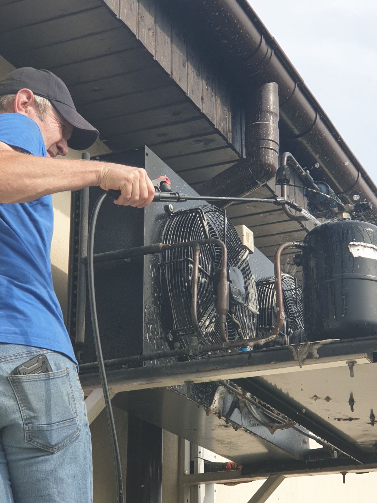 Czyszczenie myjką ciśnieniową zewnętrznej jednostki klimatyzacji na dachu budynku. Widoczny pracownik w niebieskiej koszulce i czapce z daszkiem podczas pracy.