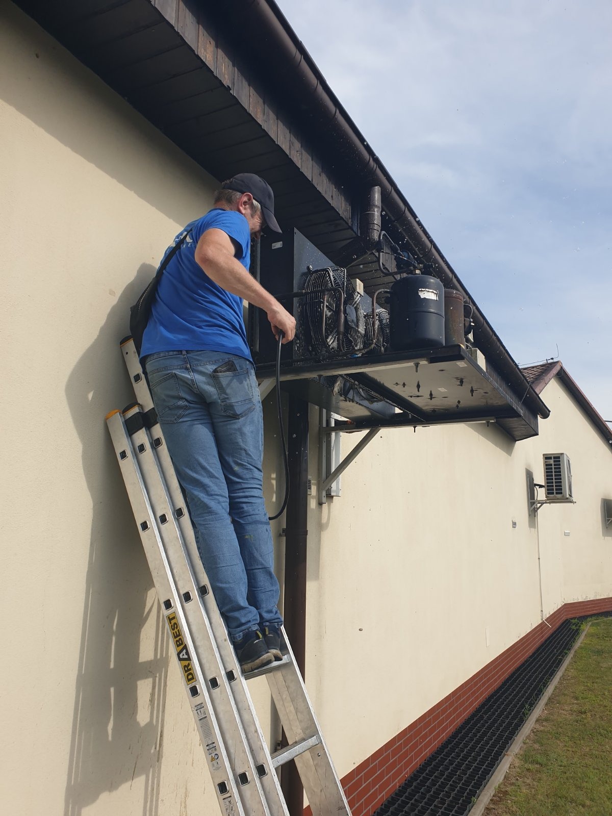 Pracownik na drabinie czyści agregat chłodniczy na zewnątrz budynku. Widoczne elementy instalacji i narzędzia pracy. Ujęcie z dołu, w tle fragment budynku.