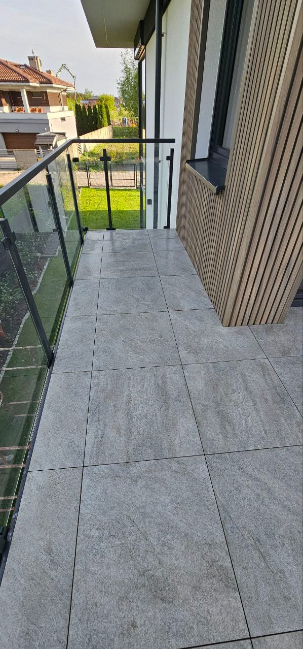 Wykończony balkon z szarą, kamienną podłogą, szklaną balustradą i drewnianą okładziną ścienną. Widok na zieleń za balustradą. Nowoczesny design.