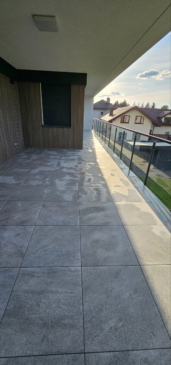 Nowoczesny balkon z szarą, kwadratową terakotą, drewnianą boazerią i szklaną balustradą. Widok na domy w tle, oświetlone popołudniowym słońcem.