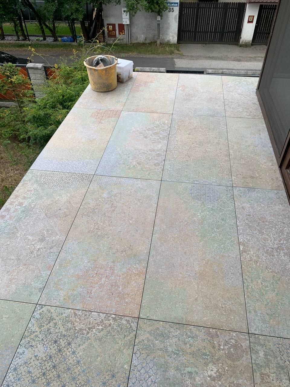 Wykończony balkon z nową, wzorzystą terakotą w odcieniach beżu i zieleni. Wiadro i akcesoria budowlane w rogu sugerują świeżo zakończony remont.