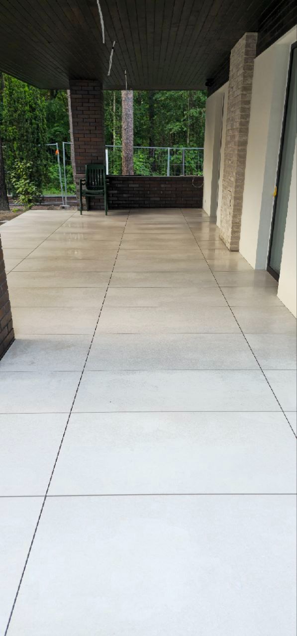 Wykończony balkon z szarymi płytami podłogowymi, ceglaną balustradą i zielonym krzesłem. Widok na zieleń w tle. Nowoczesny design z elementami naturalnymi.