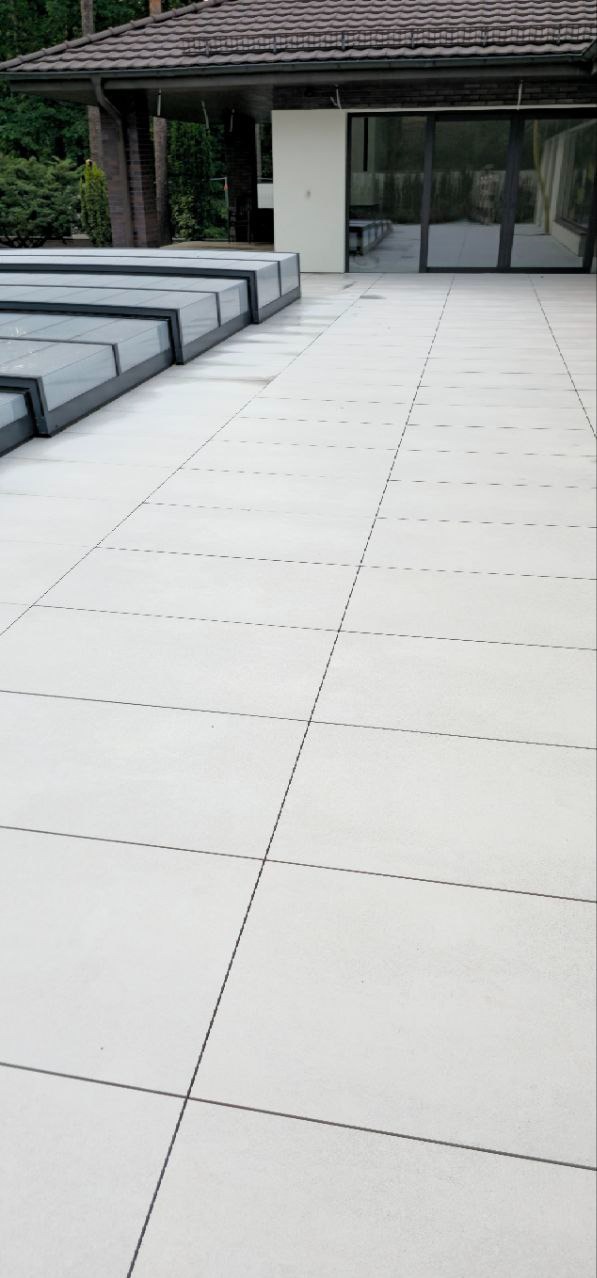 Jasne, kwadratowe płytki na tarasie z widoczną zabudową basenową i budynkiem w tle. Minimalistyczny design i precyzyjne ułożenie płytek.