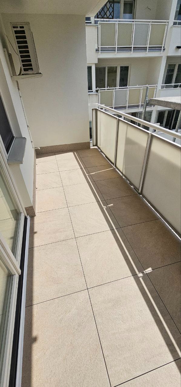 Jasny balkon z beżowymi płytkami i przeszkloną balustradą w nowoczesnym budynku. Widoczny klimatyzator na ścianie i cień balustrady na podłodze.