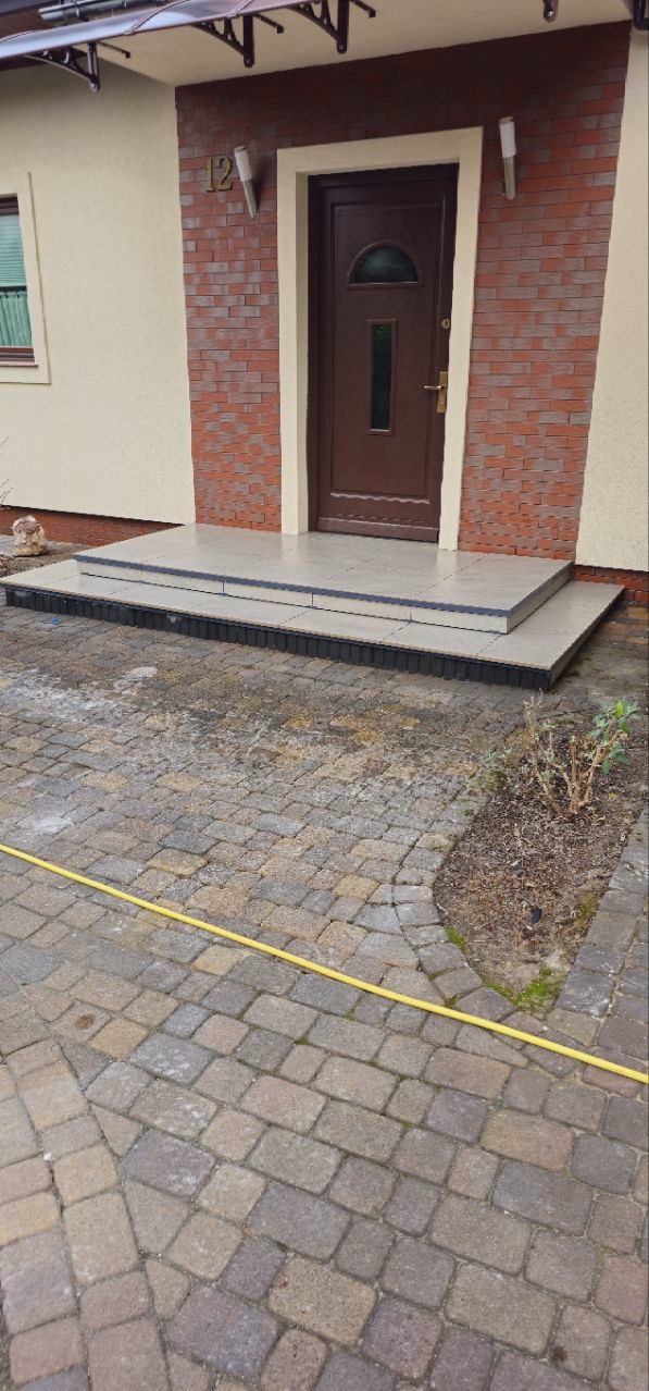 Wykończone schody wejściowe do domu z szarych płytek, z widoczną kostką brukową i fragmentem żółtego węża ogrodowego. Detal wykończenia.