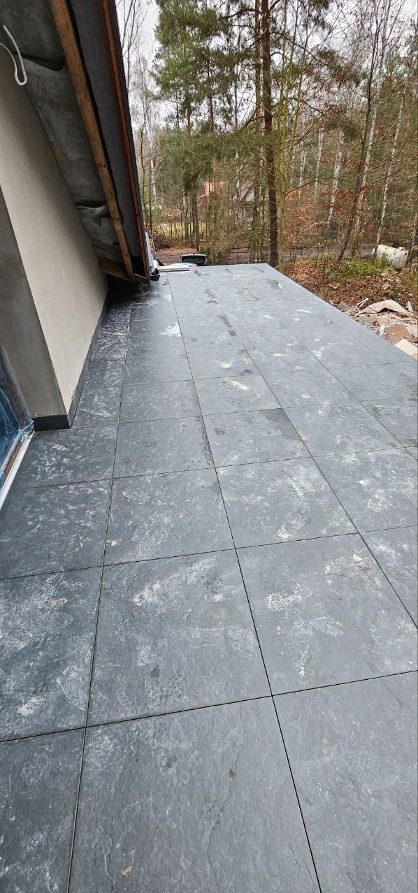 Wykończony taras z szarych, kwadratowych płytek na zewnątrz budynku, z widokiem na drzewa w tle. Widoczny fragment dachu i ściany budynku.