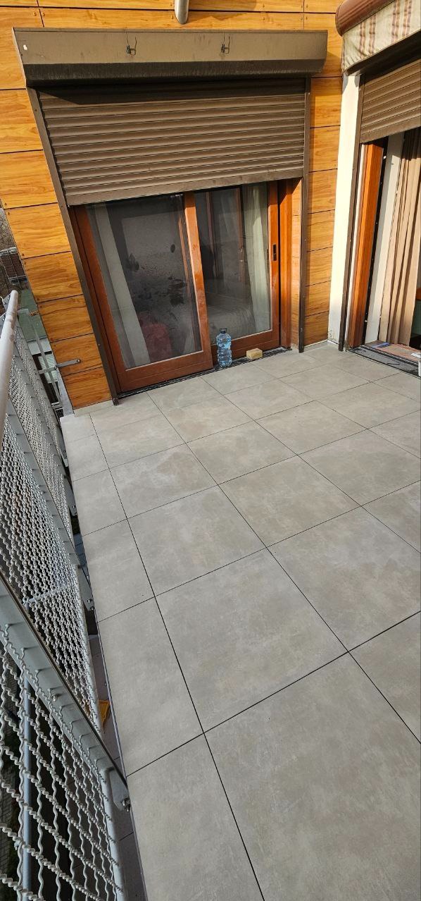 Wyłożony jasnoszarą terakotą balkon z drewnianą stolarką okienną i metalową balustradą. Widoczna butelka wody i fragment wnętrza przez okno.