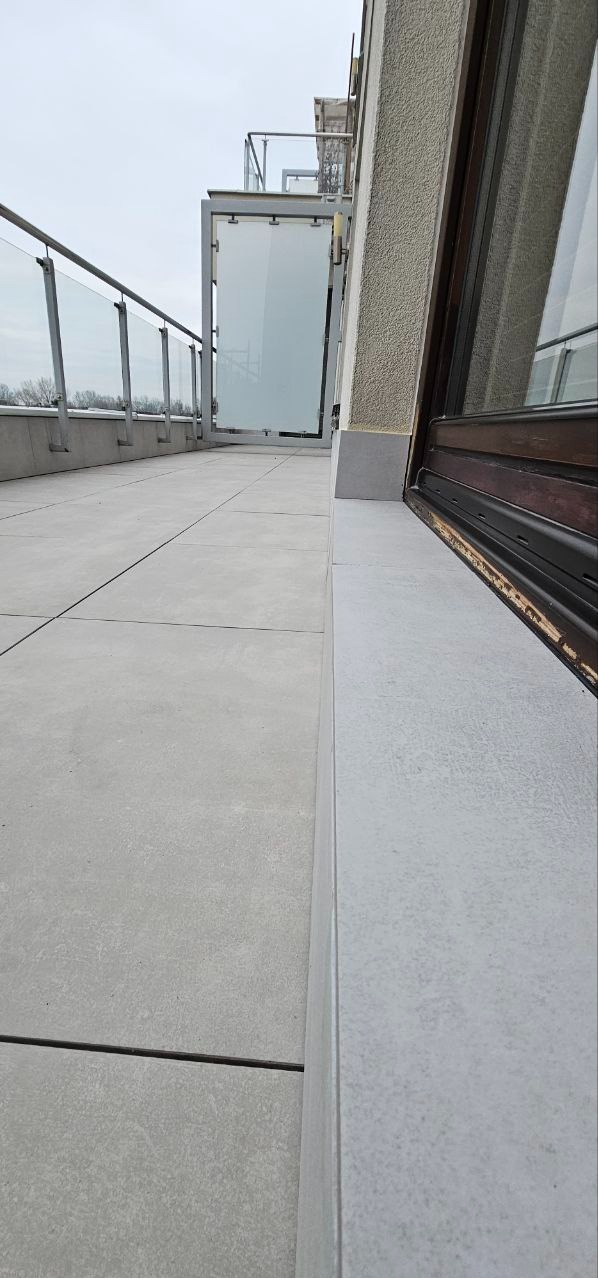 Nowoczesny balkon z szarymi płytkami, szklaną balustradą i zabudową. Minimalistyczny design, widok z boku, neutralna kolorystyka.