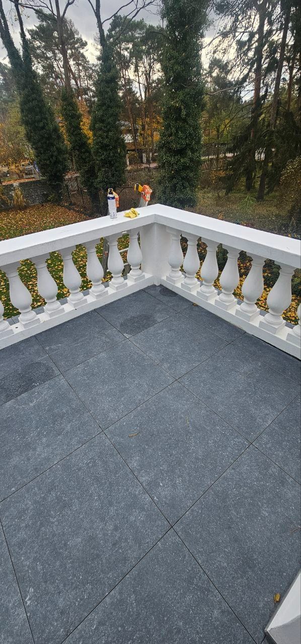 Wykończony balkon z szarą terakotą i białą balustradą. W tle zieleń drzew. Na balustradzie puszka farby i ściereczka, sugerujące świeżo zakończone prace malarskie.