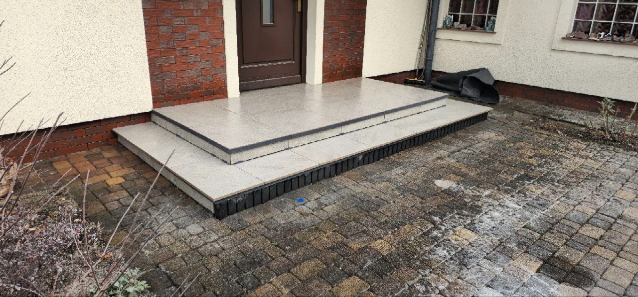 Wykończone płytkami schody wejściowe do domu z brązowymi drzwiami i ceglaną elewacją. Kostka brukowa na podjeździe. Widok z dołu.