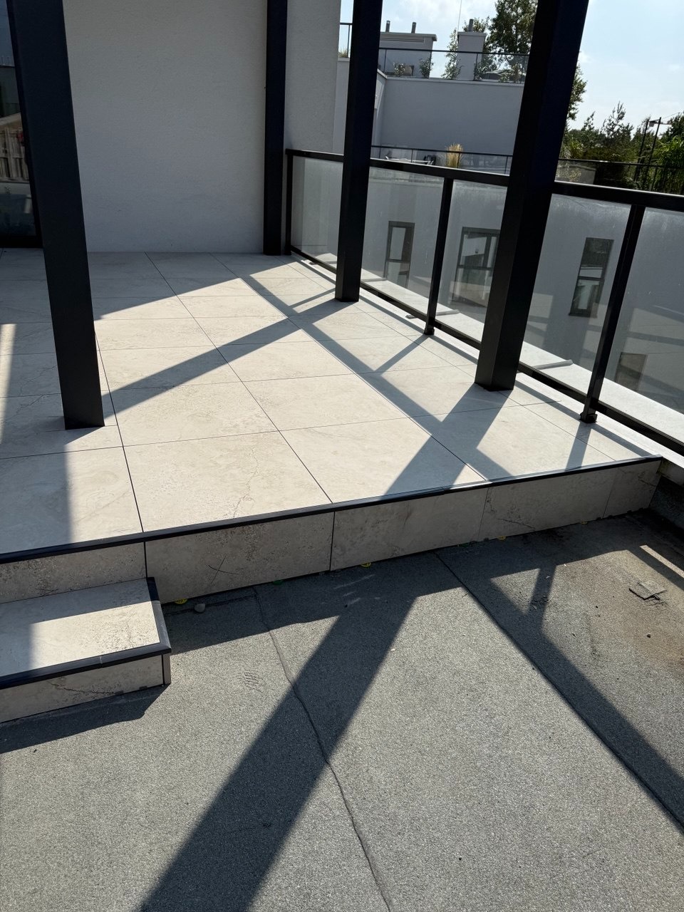 Wykończony balkon z jasnymi płytkami, czarnymi słupkami i szklanymi balustradami. Widoczne cienie słoneczne na podłodze i fragment dachu budynku w tle.
