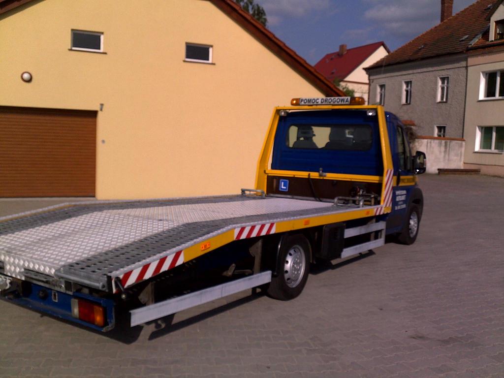 Niebiesko-żółta autolaweta z napisem 'POMOC DROGOWA' na dachu, zaparkowana na kostce brukowej przed budynkami.