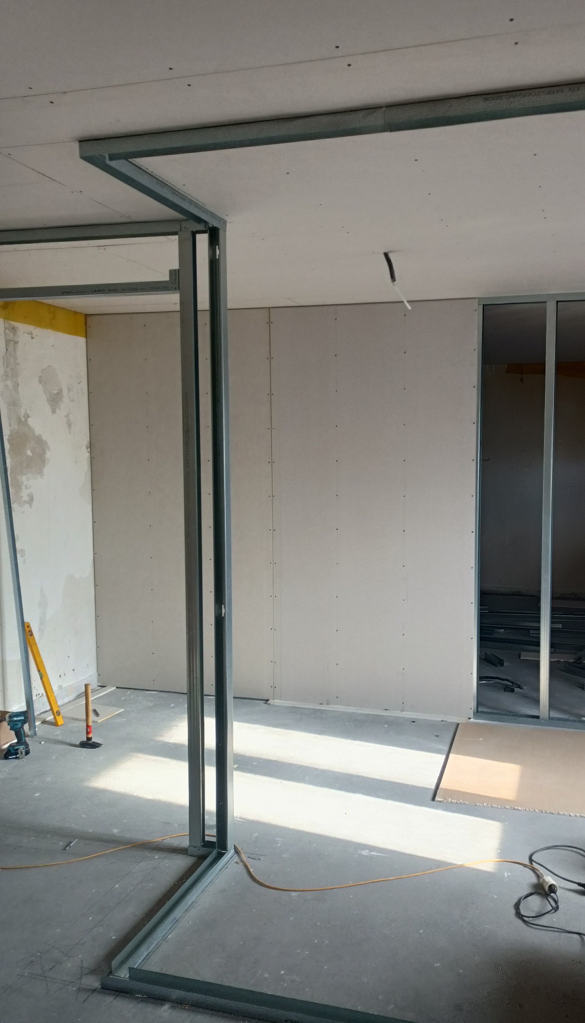 Montaż metalowej konstrukcji pod płyty gipsowe na ścianie i suficie. Widoczne narzędzia i fragmenty płyt. Wnętrze w trakcie prac wykończeniowych.