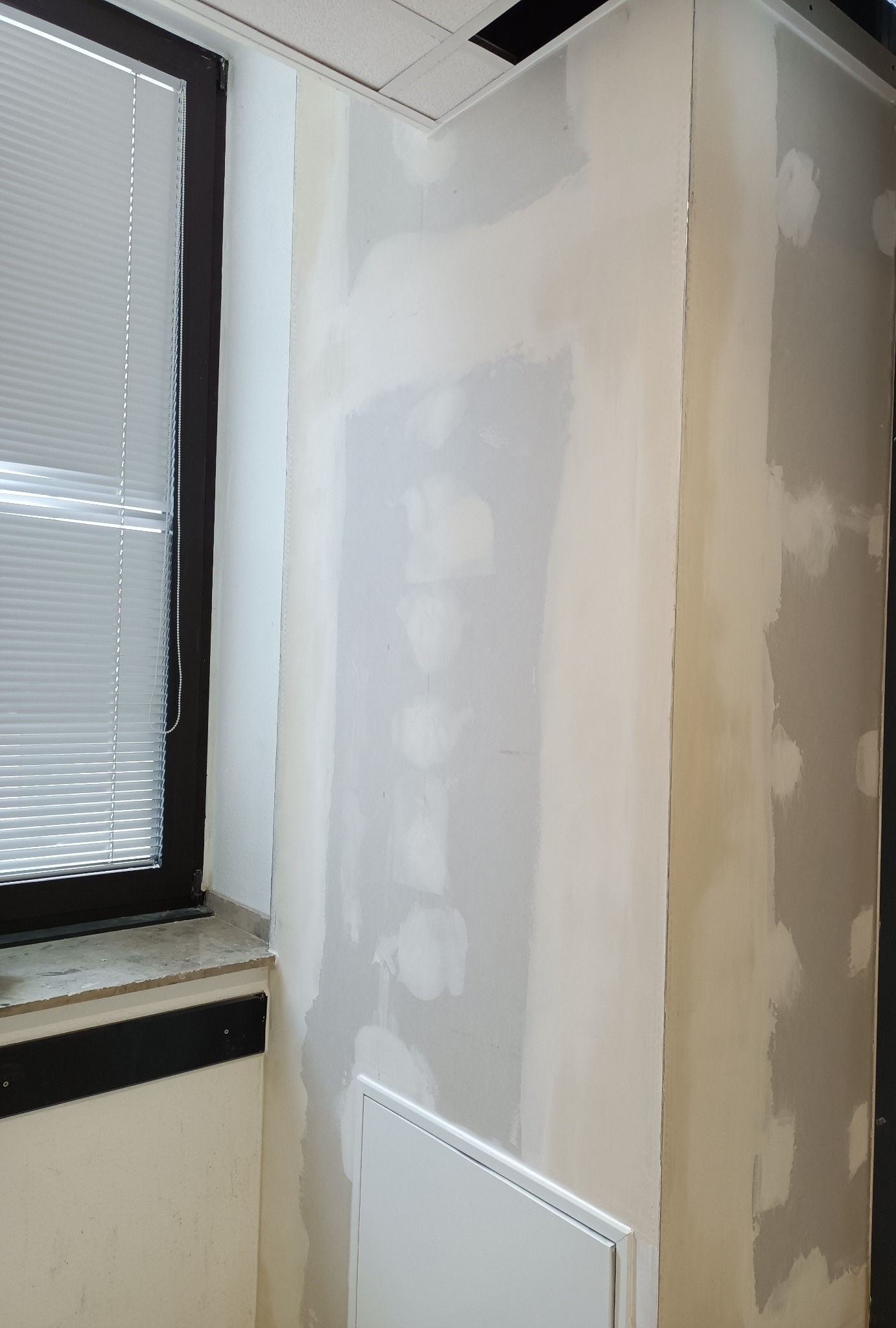 Ściana w trakcie szpachlowania, widoczne nierówności i miejsca po łatach. Okno z żaluzjami w tle. Etap prac wykończeniowych w pomieszczeniu biurowym.