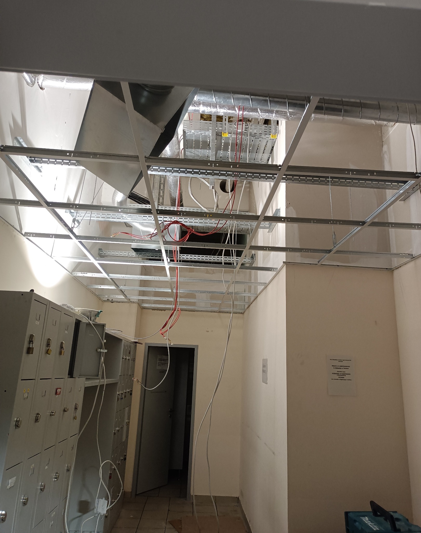 Montaż sufitu podwieszanego z widoczną instalacją wentylacyjną i elektryczną w pomieszczeniu z szafkami. Widoczne kable i elementy konstrukcyjne.