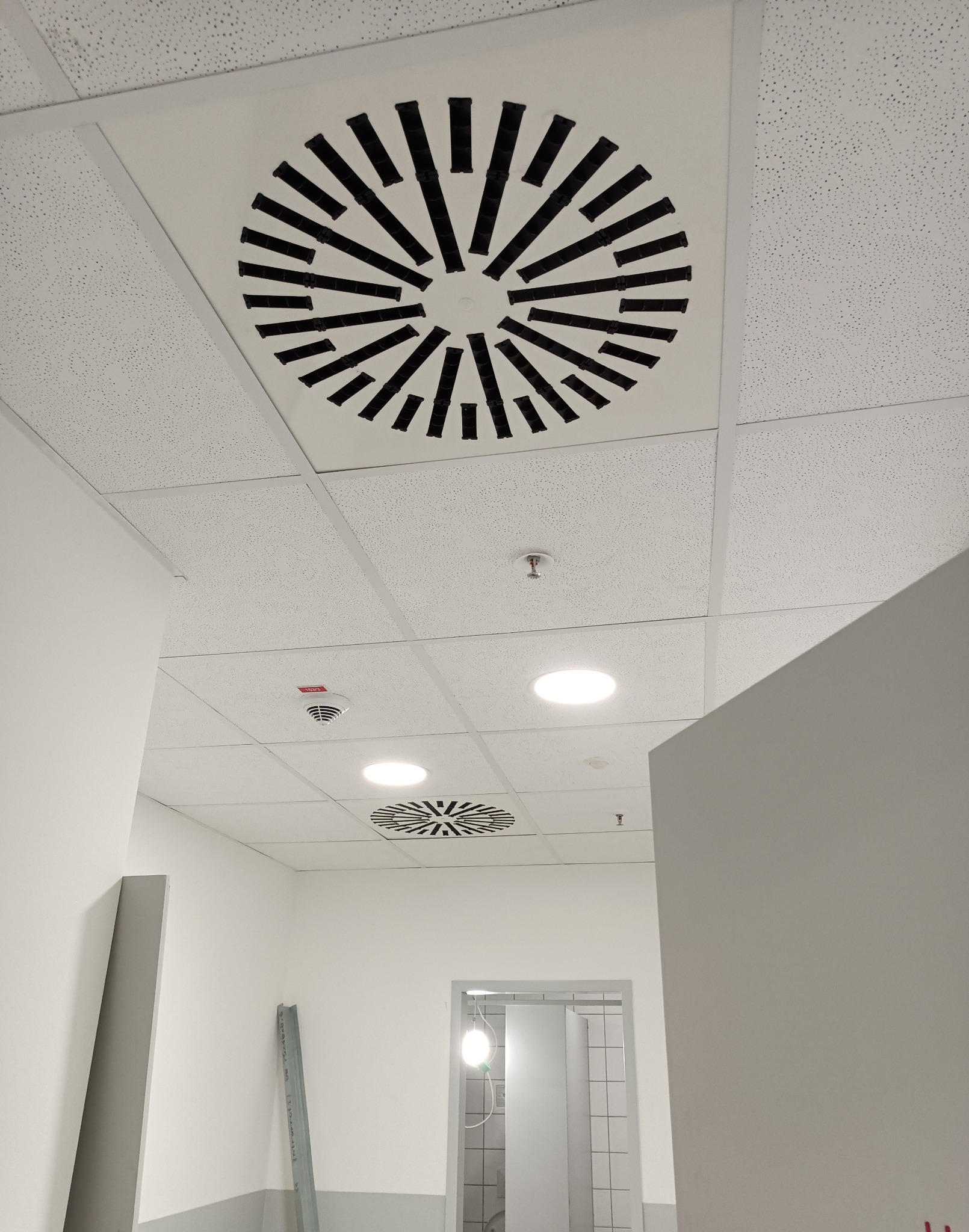 Biały sufit podwieszany z perforacją i okrągłymi kratkami wentylacyjnymi z czarnymi elementami. Widoczne oświetlenie i fragment korytarza z drzwiami.