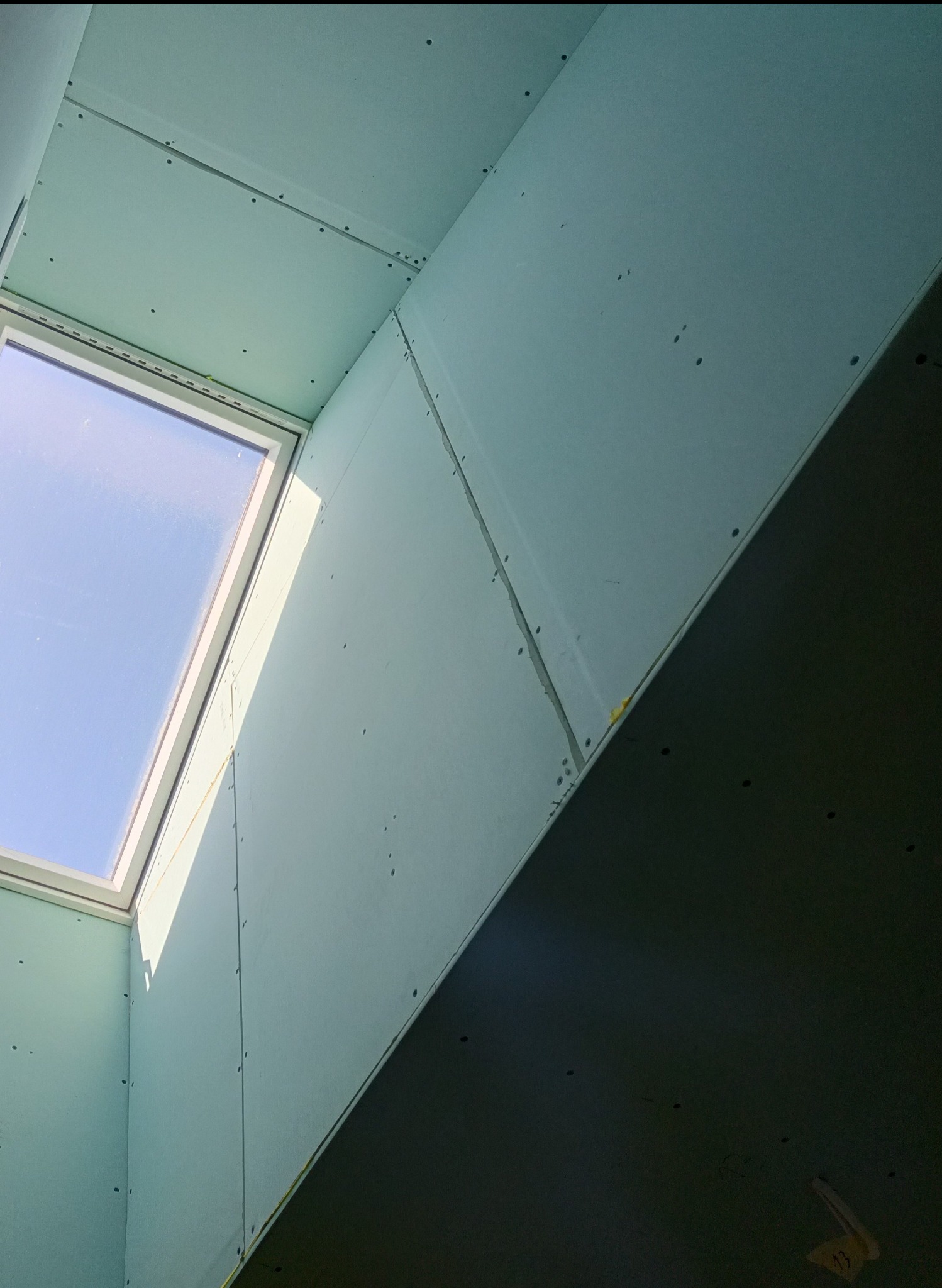 Ściana i sufit z zamontowanymi płytami, widoczne łączenia i wkręty. W tle okno dachowe z widokiem na niebo. Prace wykończeniowe w nowym domu.