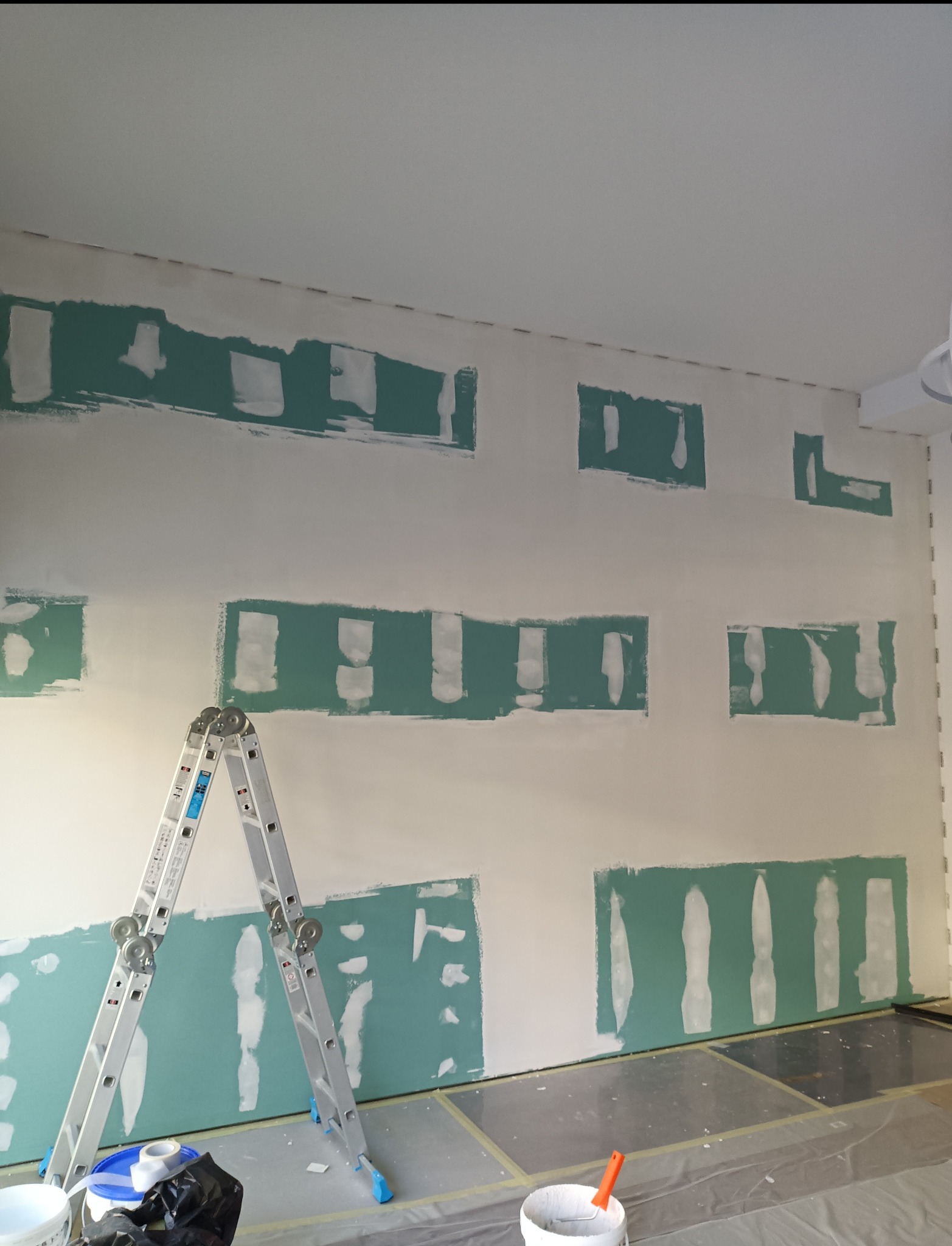 Ściana z nałożonymi łatami gipsowymi, przygotowana do dalszych prac wykończeniowych. Widoczna drabina i akcesoria malarskie.