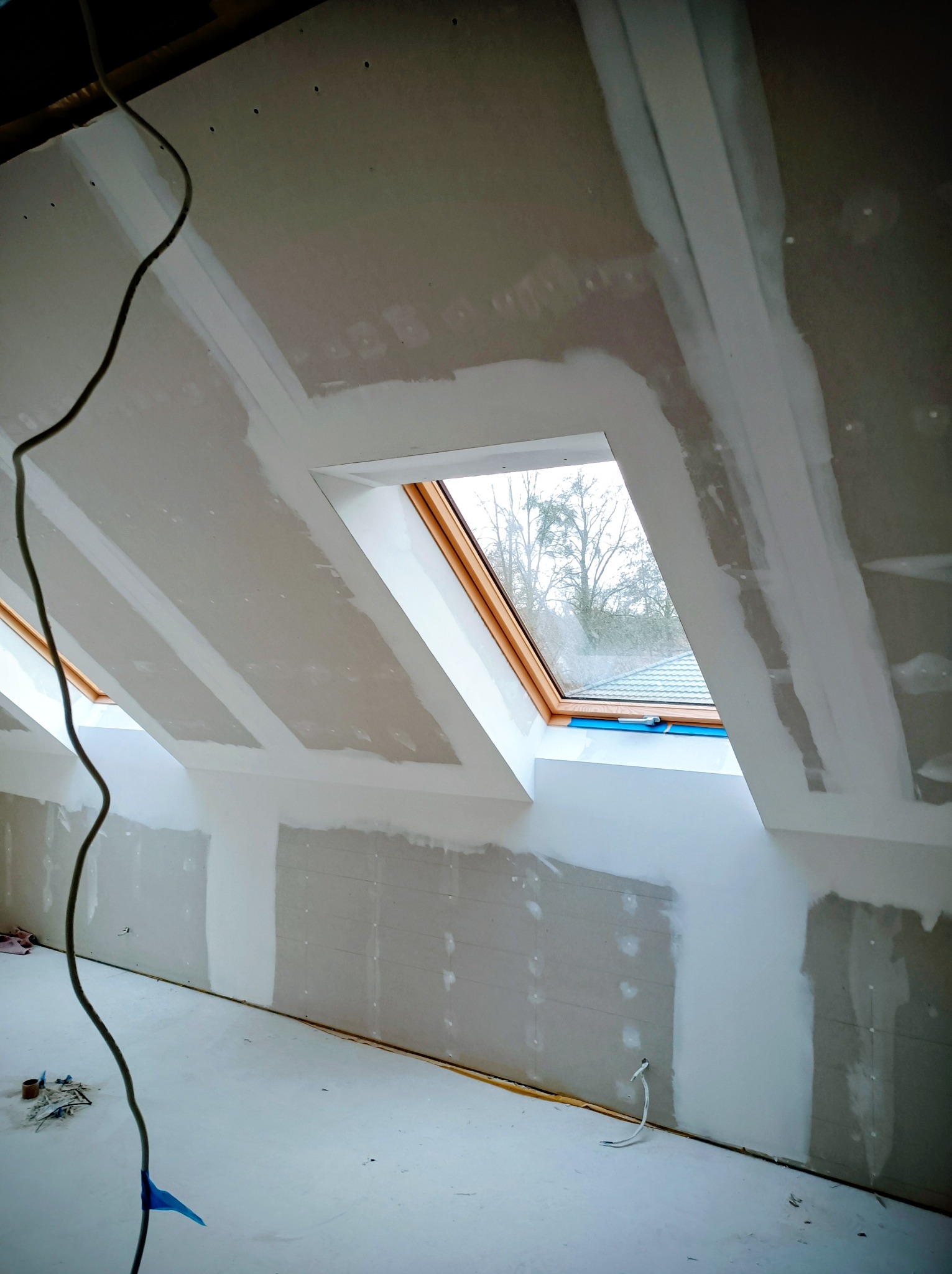 Poddasze z oknami dachowymi w trakcie prac wykończeniowych. Widoczne płyty gipsowo-kartonowe i wstępnie nałożona gładź. Kabel elektryczny zwisa z sufitu.