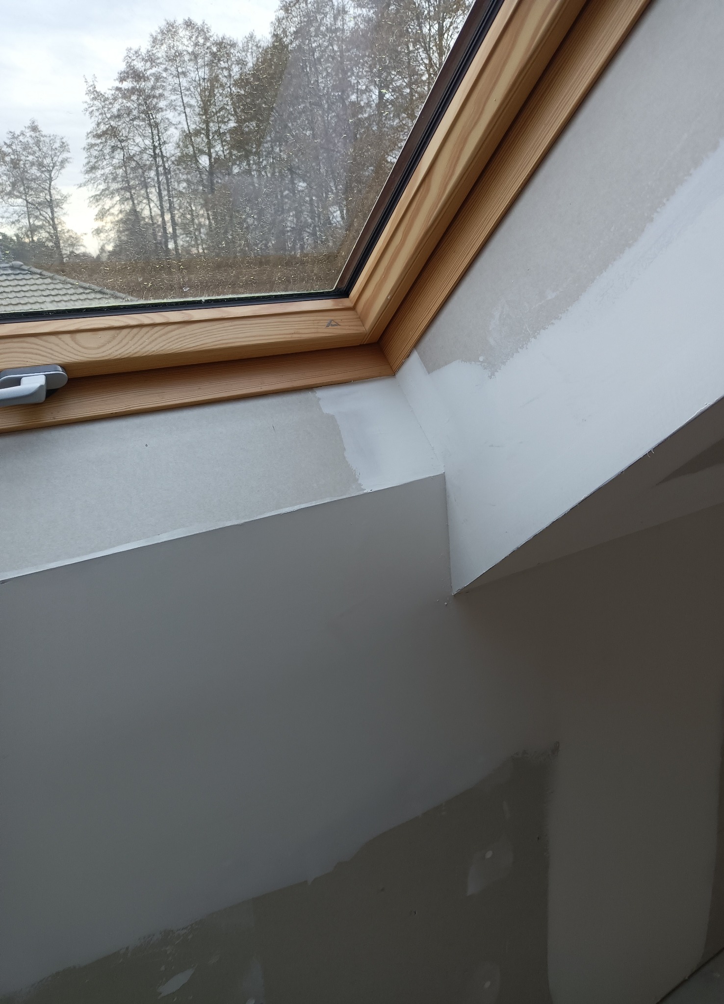 Ściana pod skosem z oknem dachowym w trakcie prac wykończeniowych, częściowo pokryta gładzią. Widoczne drewniane ramy okna i fragment krajobrazu za szybą.
