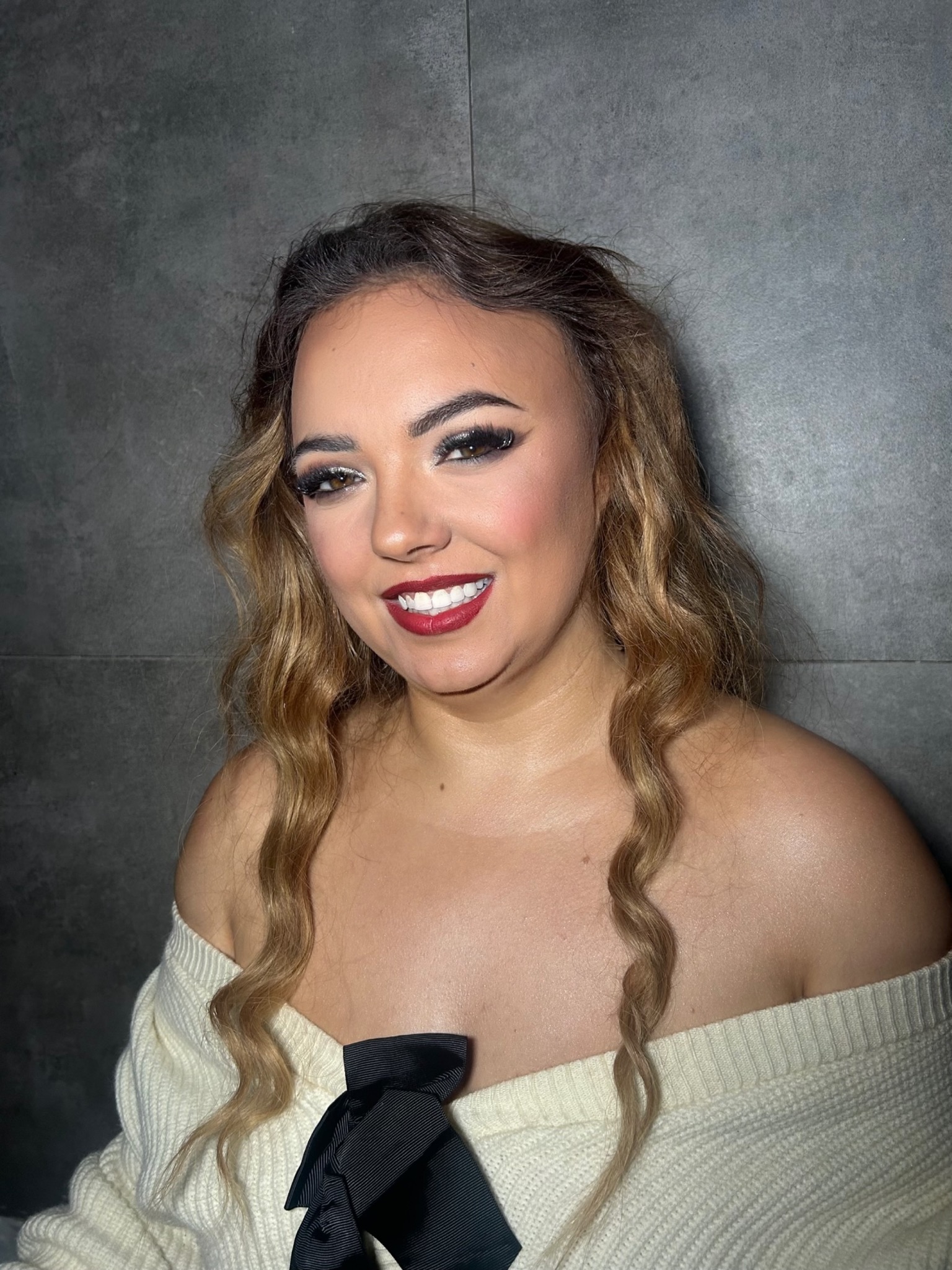 Uśmiechnięta blondynka z wyrazistym makijażem oczu i czerwonymi ustami, ubrana w biały sweter z czarną kokardą na tle szarej ściany.