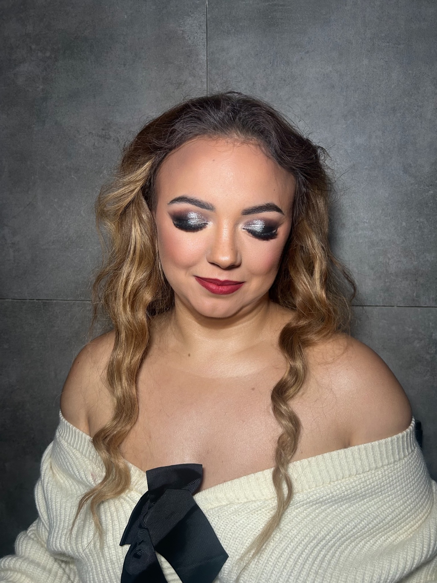 Portret kobiety z zamkniętymi oczami, prezentującej wieczorowy makijaż z brokatem na powiekach i ciemną szminką, w białym swetrze z kokardą na tle szarej ściany.