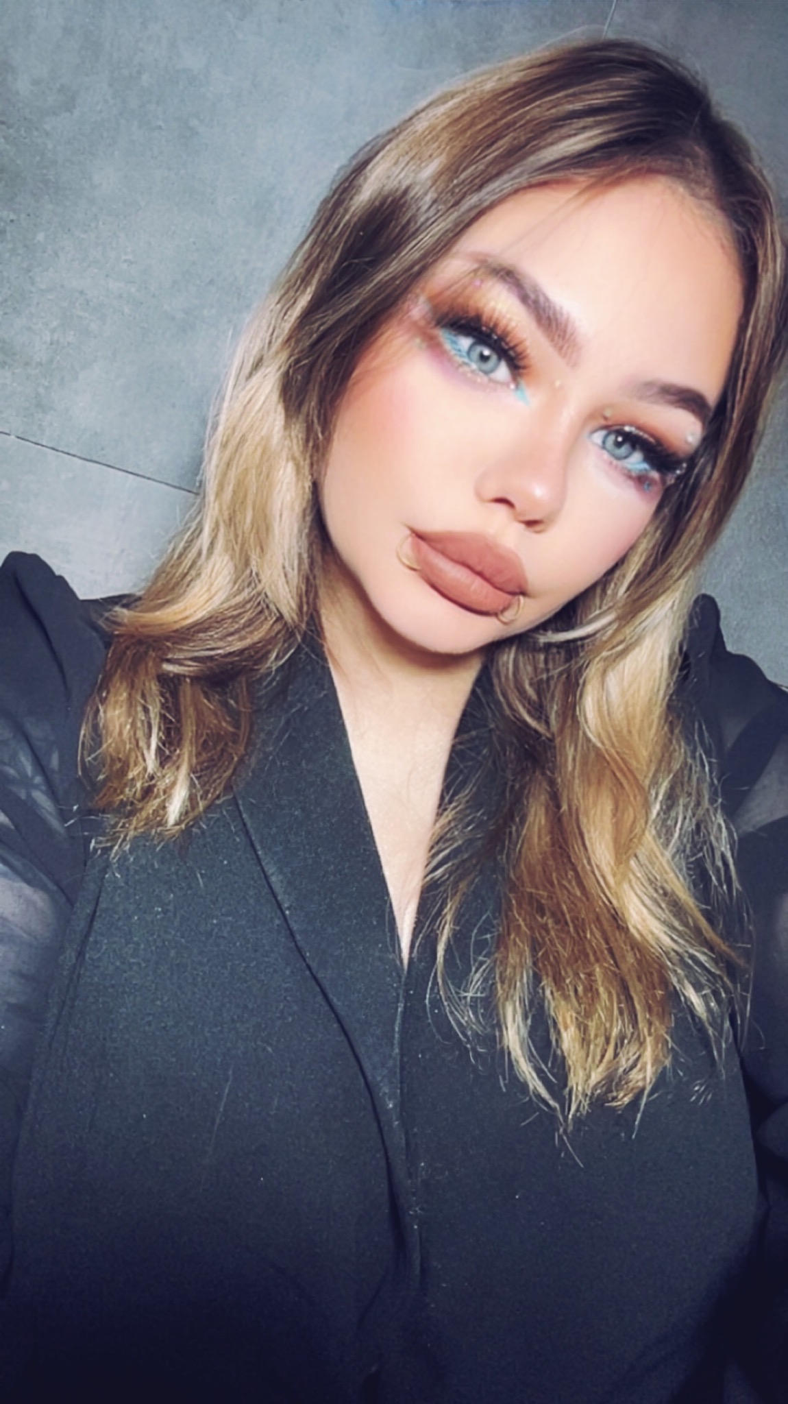 Portret kobiety z intensywnym makijażem oczu w odcieniach błękitu i złota, z piercingiem w wardze, ubrana w czarną marynarkę na tle szarej ściany.