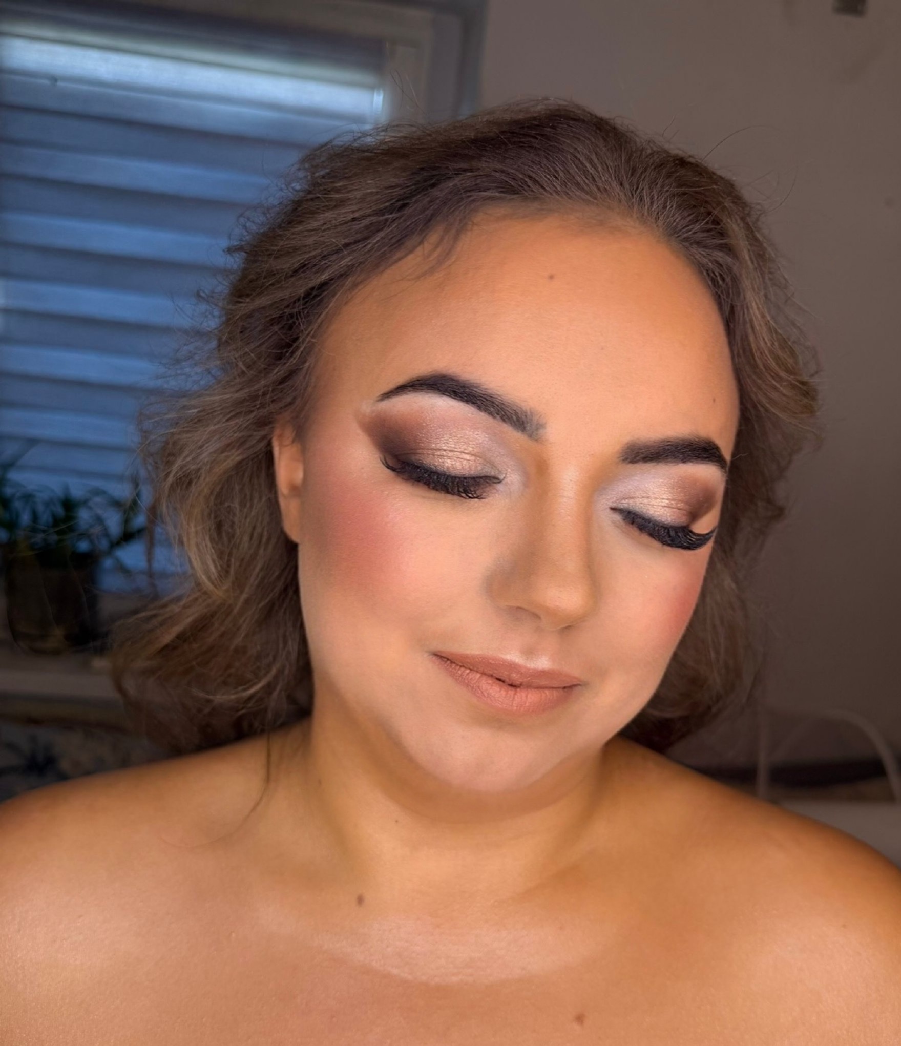 Portret kobiety z zamkniętymi oczami, prezentujący makijaż w odcieniach brązu i beżu, z wyraźnie podkreślonymi rzęsami i policzkami. Subtelne fale włosów.