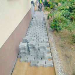 UR-BUD PIOTR URBAN - Układanie szarej kostki brukowej typu 'ażur' wzdłuż ściany budynku. Część kostki ułożona, część w stosie. Widoczny pracownik z taczką w tle.