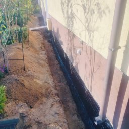 UR-BUD PIOTR URBAN - Wykop fundamentowy wzdłuż ściany budynku, zabezpieczony czarną folią hydroizolacyjną. Widoczny rów z ziemią i fragment systemu rynnowego.