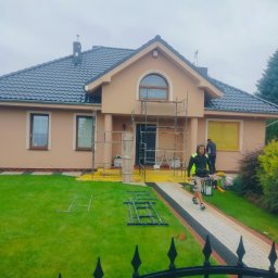 UR-BUD PIOTR URBAN - Dwukondygnacyjny dom z beżową elewacją w trakcie prac wykończeniowych. Widoczne rusztowania, pracownicy i zabezpieczenia okien folią. Ujęcie zza ozdobnego, metalowego płotu.