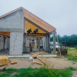 UR-BUD PIOTR URBAN - Wznoszenie domu z białych bloczków: murarze montują drewnianą konstrukcję dachu na surowych ścianach. Widoczne rusztowania, materiały budowlane i otoczenie placu budowy.