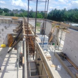 UR-BUD PIOTR URBAN - Zbrojenie fundamentów budynku z bloczków betonowych, widoczne szalunki i elementy konstrukcyjne. W tle zieleń drzew i błękitne niebo z chmurami.