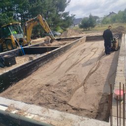 UR-BUD PIOTR URBAN - Fundamenty domu w trakcie budowy: wykop z izolacją, ubijanie piasku zagęszczarką, widoczny sprzęt budowlany i zbrojenie. Prace ziemne w słoneczny dzień.