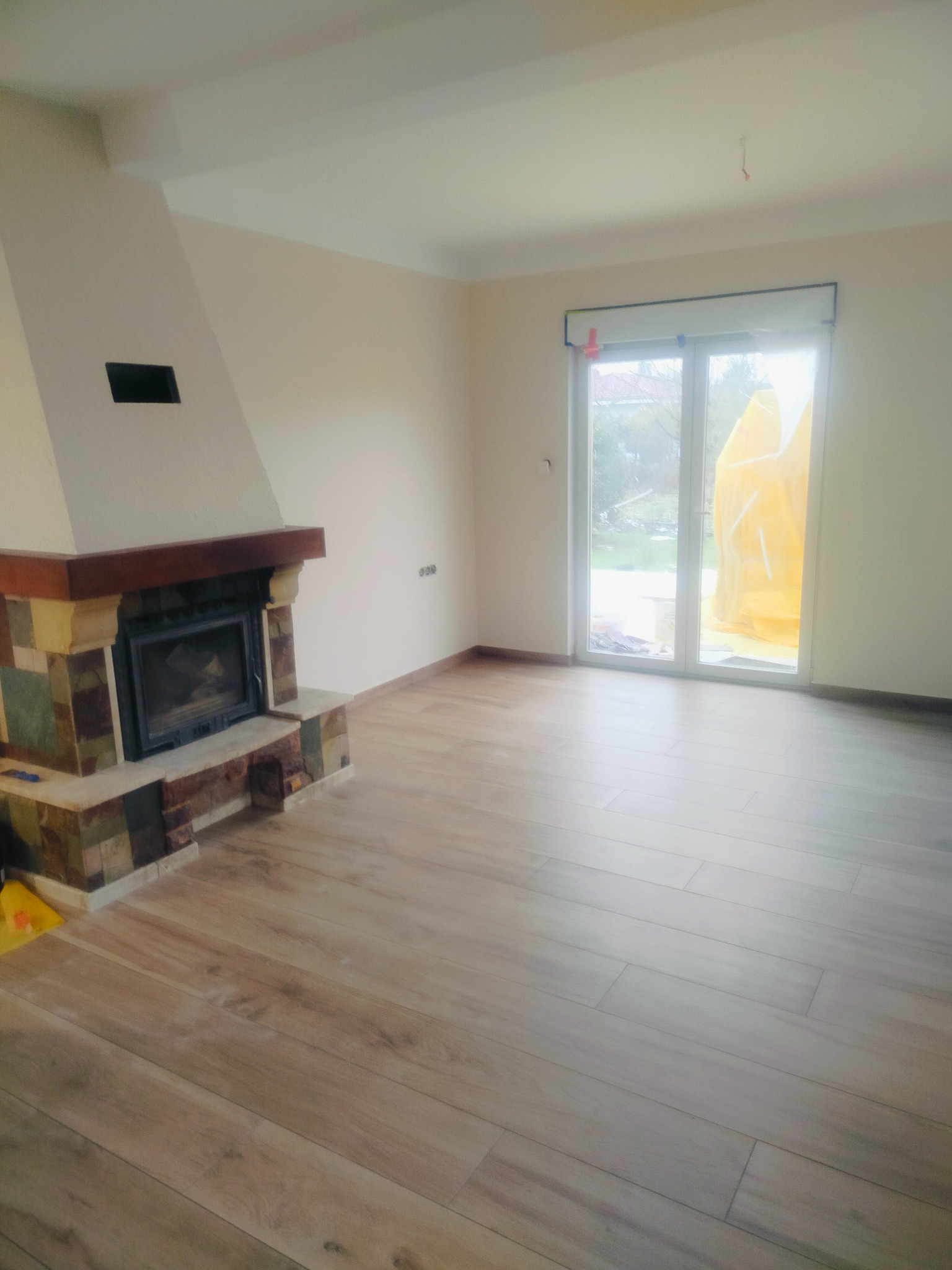 Jasne wnętrze z nową podłogą imitującą drewno i kominkiem z okładziną z kamienia. Widoczne drzwi balkonowe z żółtą folią ochronną.