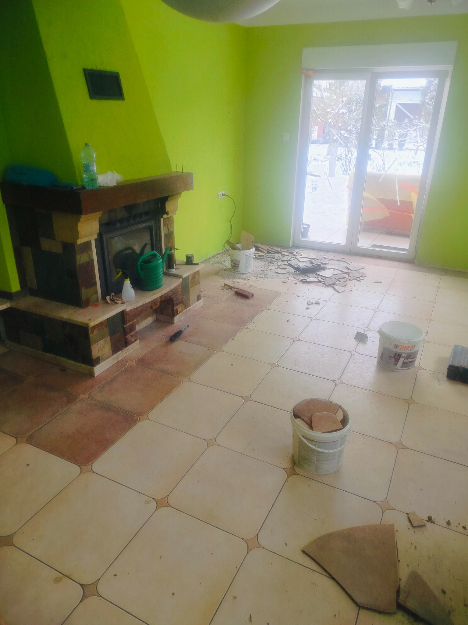 Remont podłogi z kominkiem w jaskrawozielonym pokoju. Częściowo zerwane płytki, widoczne narzędzia i materiały budowlane. Prace w toku, bałagan na podłodze.