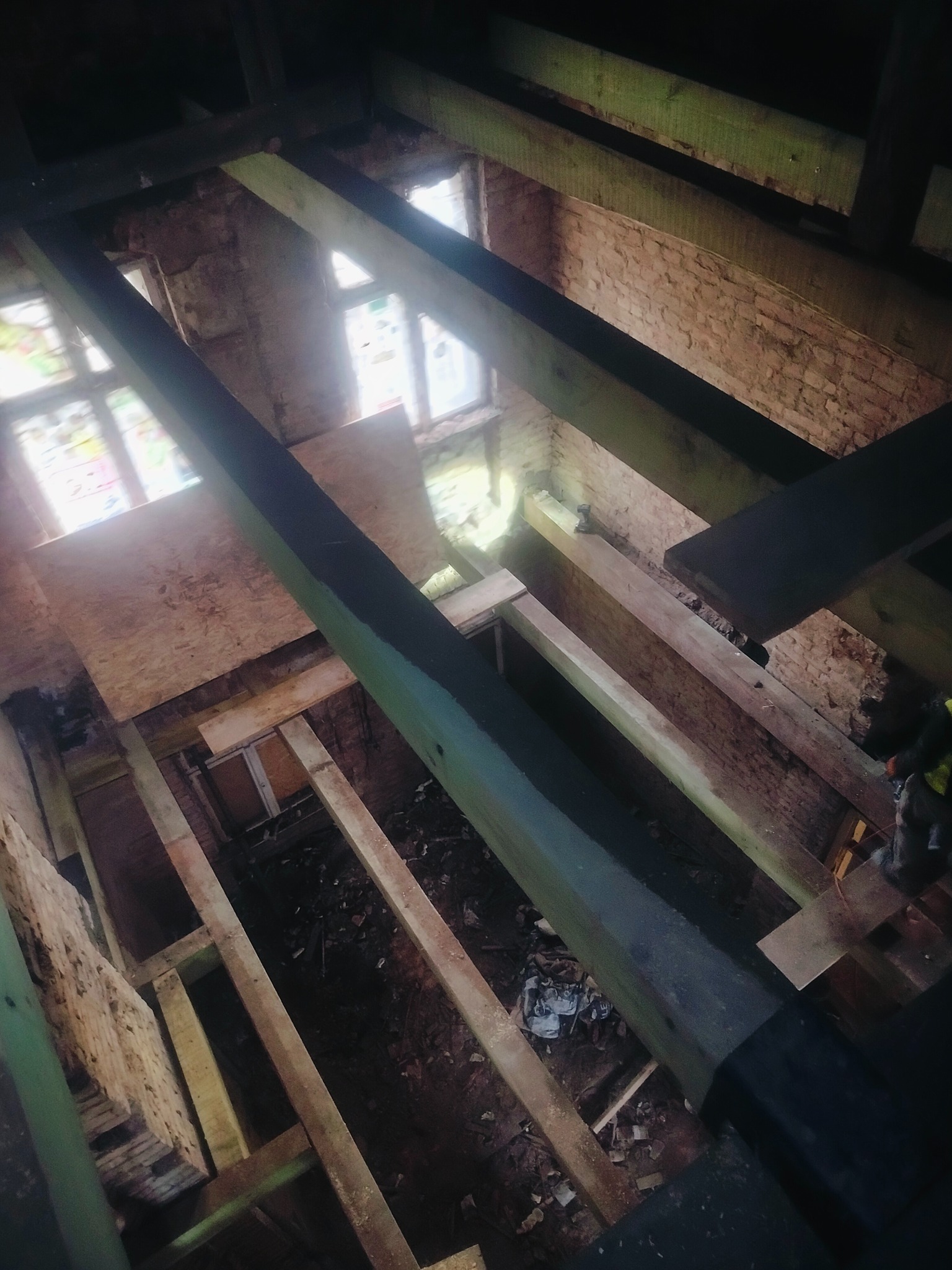 Wnętrze remontowanego poddasza z drewnianą konstrukcją stropu, widoczne okna i fragment muru. Prace remontowe w toku, surowe drewno i elementy konstrukcyjne.