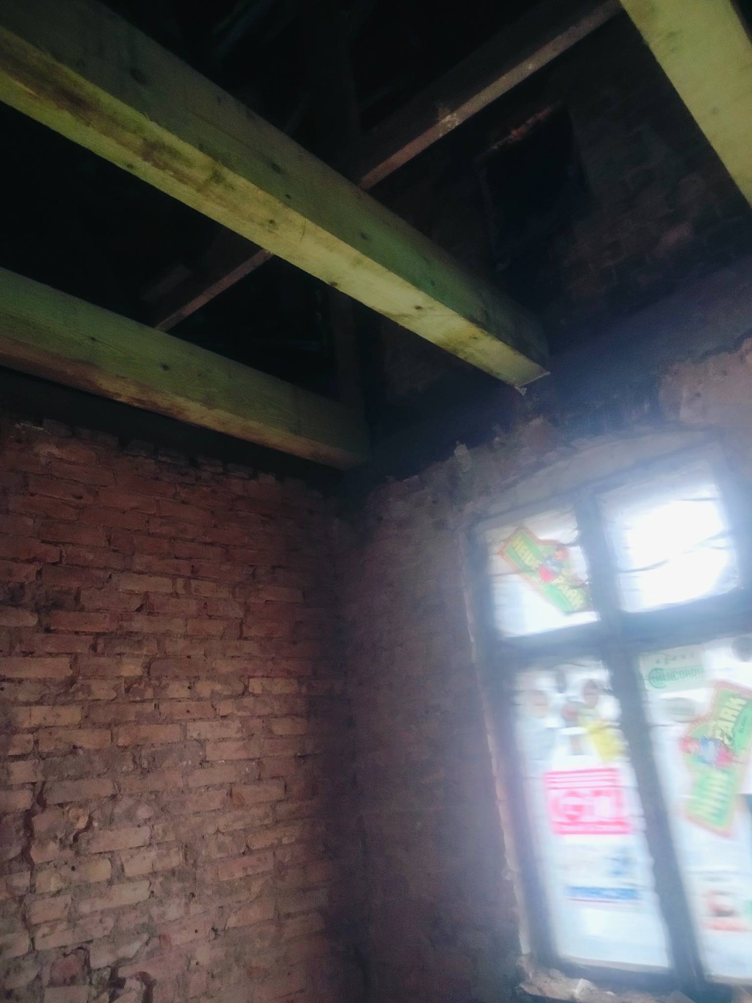 Wnętrze starego budynku z ceglaną ścianą, drewnianymi belkami stropowymi i oknem z naklejonymi plakatami. Widoczne ślady zawilgocenia i zaniedbania.