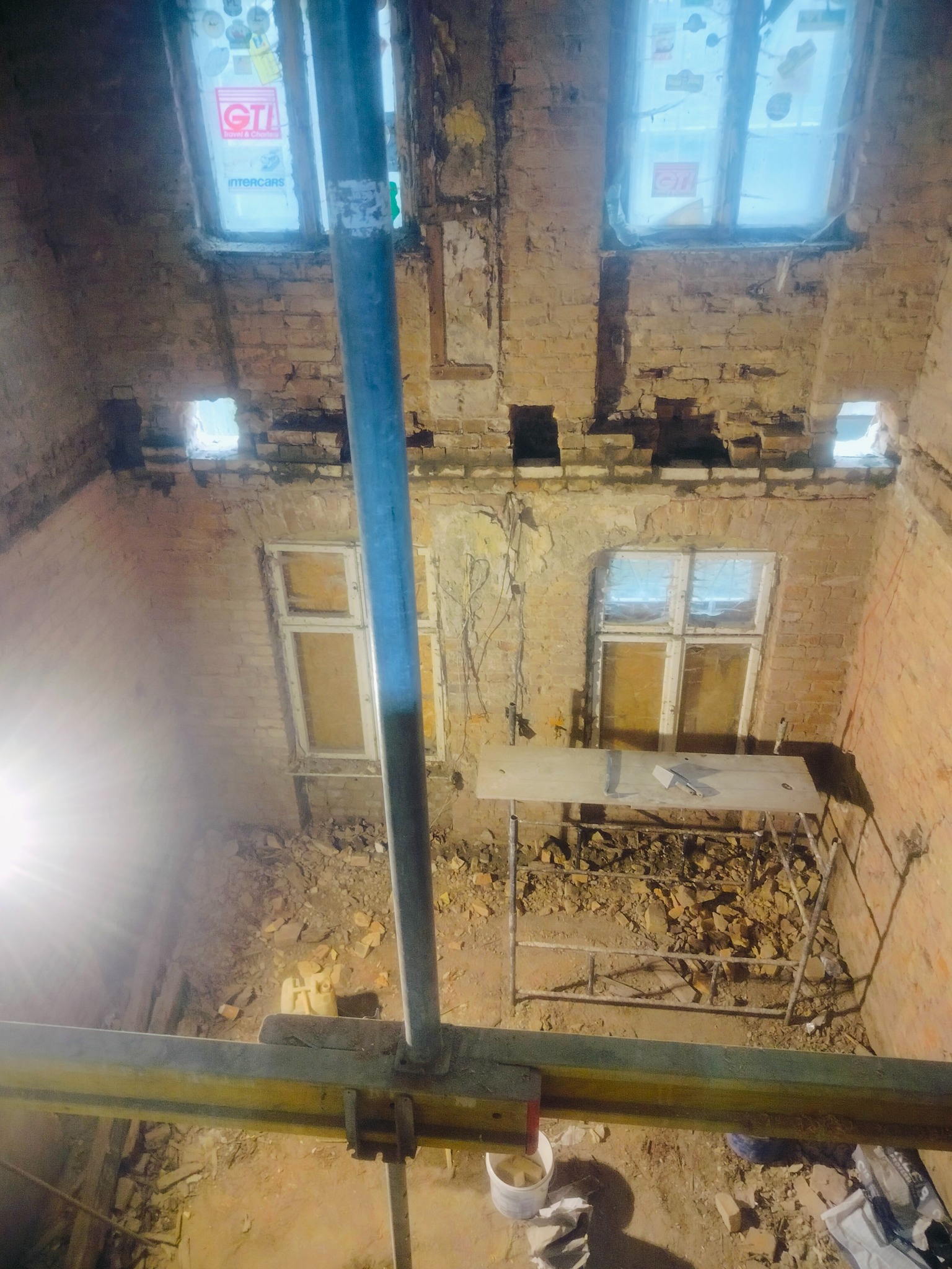 Wnętrze remontowanego budynku z ceglanymi ścianami, widoczne rusztowania i okna zabezpieczone folią. Fragmenty ścian usunięte, przygotowanie do dalszych prac.
