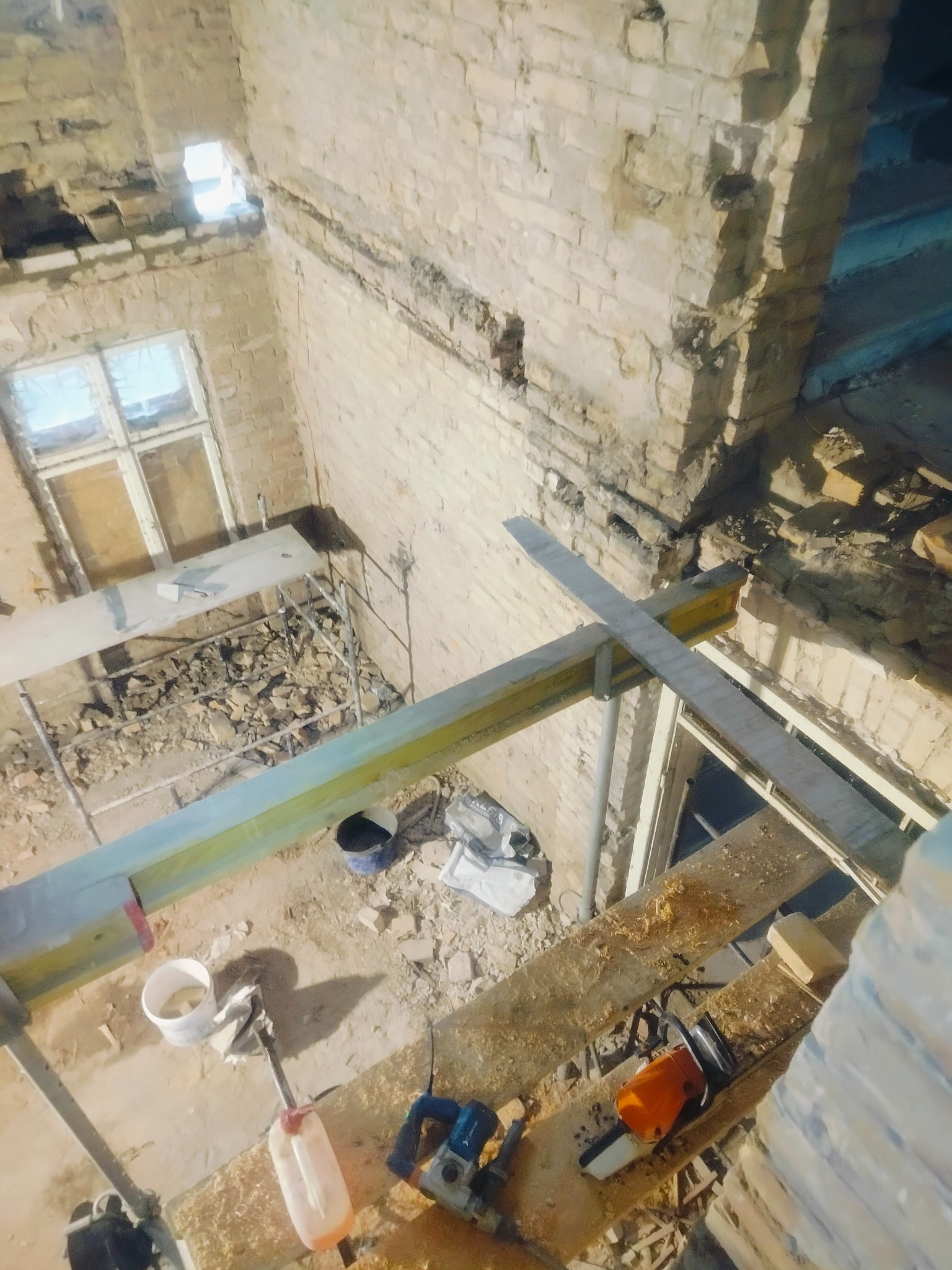 Wnętrze remontowanego budynku z cegły, widok z góry na rusztowania, narzędzia i gruz. Widoczne okno i fragmenty ścian w trakcie prac murarskich.