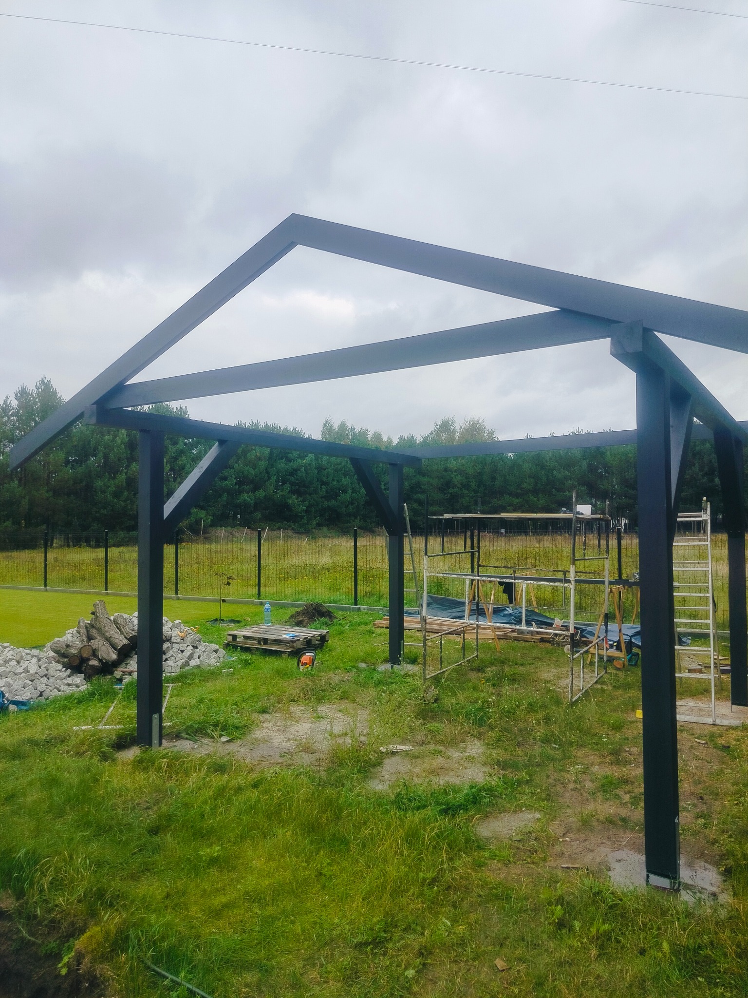 Konstrukcja z ciemnego drewna w ogrodzie, prawdopodobnie altana lub pergola w trakcie budowy. Widoczne rusztowania i materiały budowlane. Tło stanowią drzewa i pochmurne niebo.
