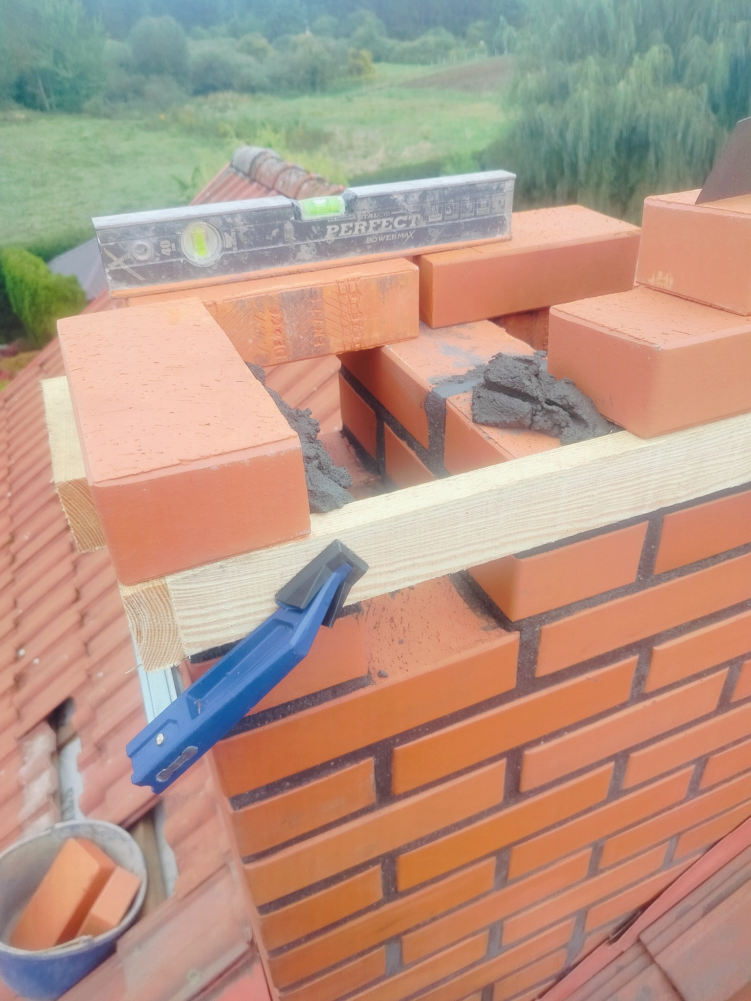 Murowanie komina z cegły ceramicznej na dachu, poziomica, drewniane podpory, kielnia, wiadro z cegłami. Widok z góry na etap budowy komina.