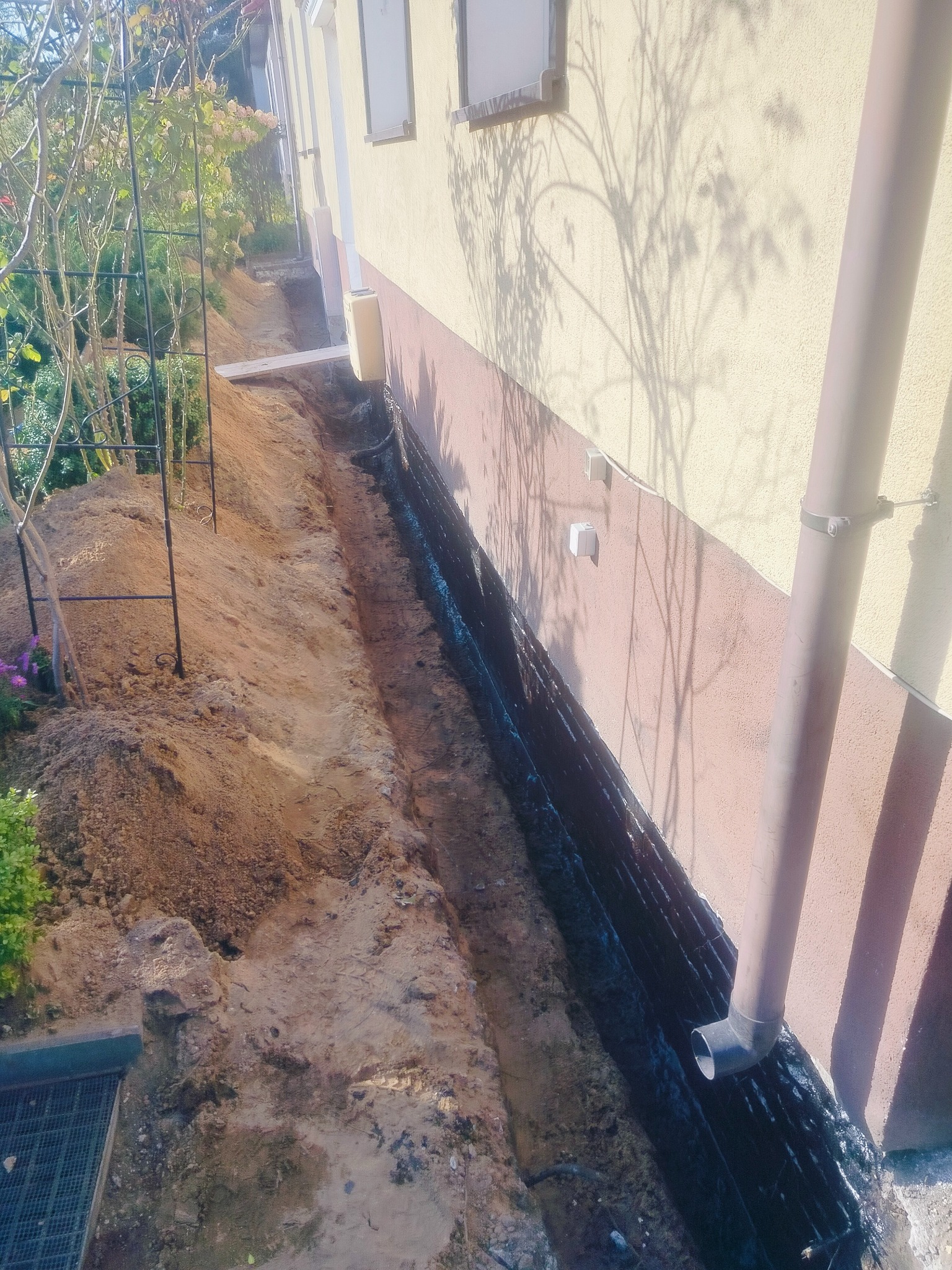 Wykop fundamentowy wzdłuż ściany budynku, zabezpieczony czarną folią hydroizolacyjną. Widoczny rów z ziemią i fragment systemu rynnowego.