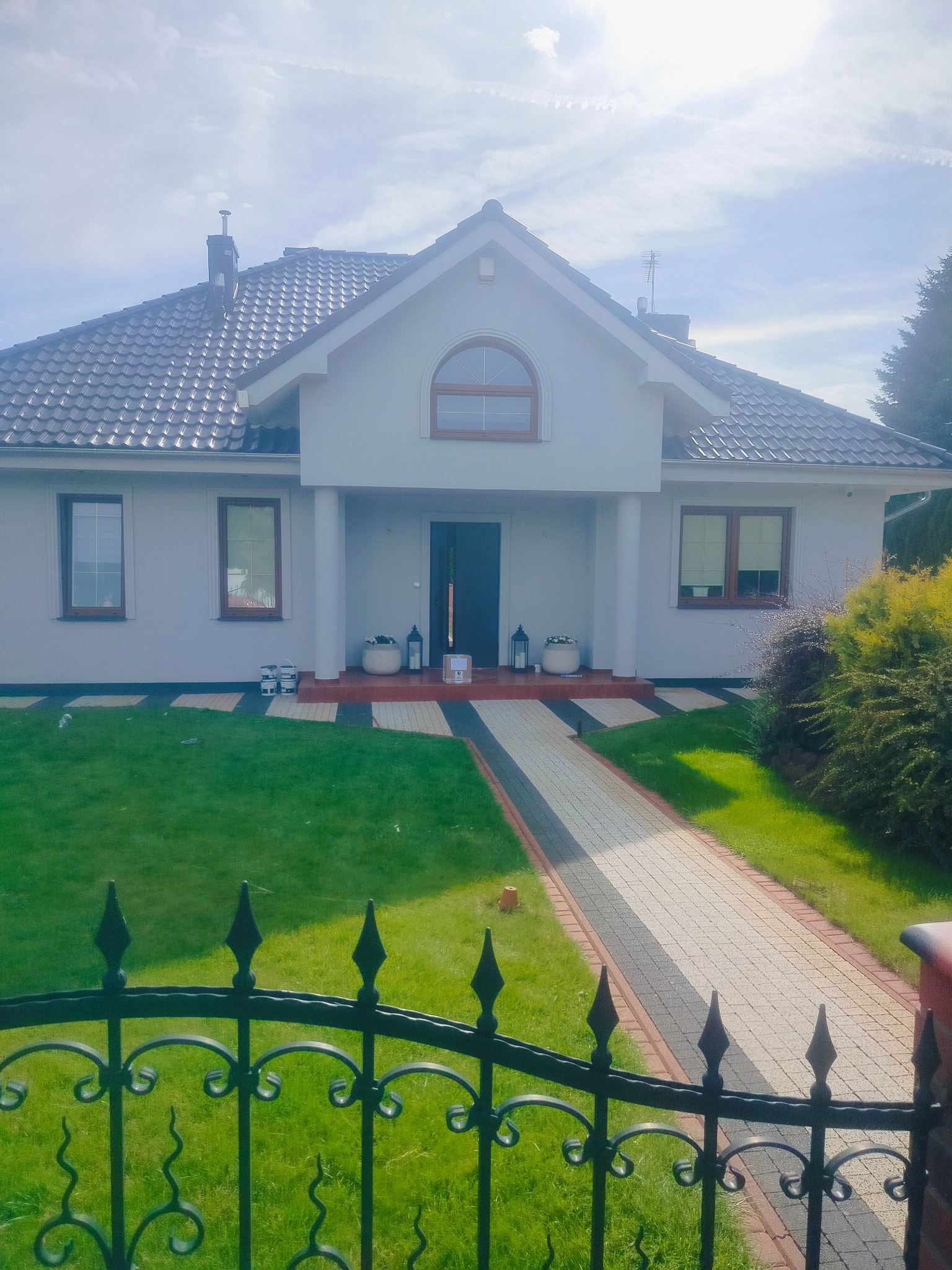 Elegancki dom jednorodzinny w Gorzowie Wielkopolskim po remoncie elewacji, widoczny zza ozdobnego, kutego ogrodzenia. Świeżo pomalowana fasada w jasnym odcieniu.