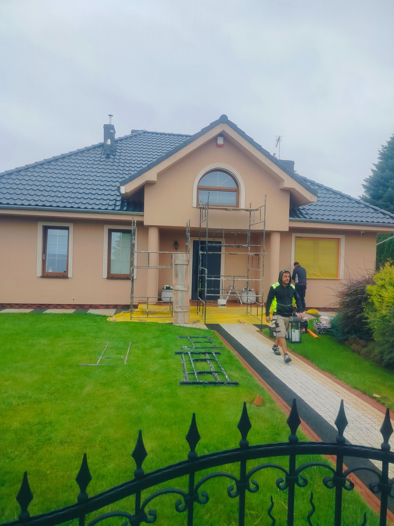 Dwukondygnacyjny dom z beżową elewacją w trakcie prac wykończeniowych. Widoczne rusztowania, pracownicy i zabezpieczenia okien folią. Ujęcie zza ozdobnego, metalowego płotu.