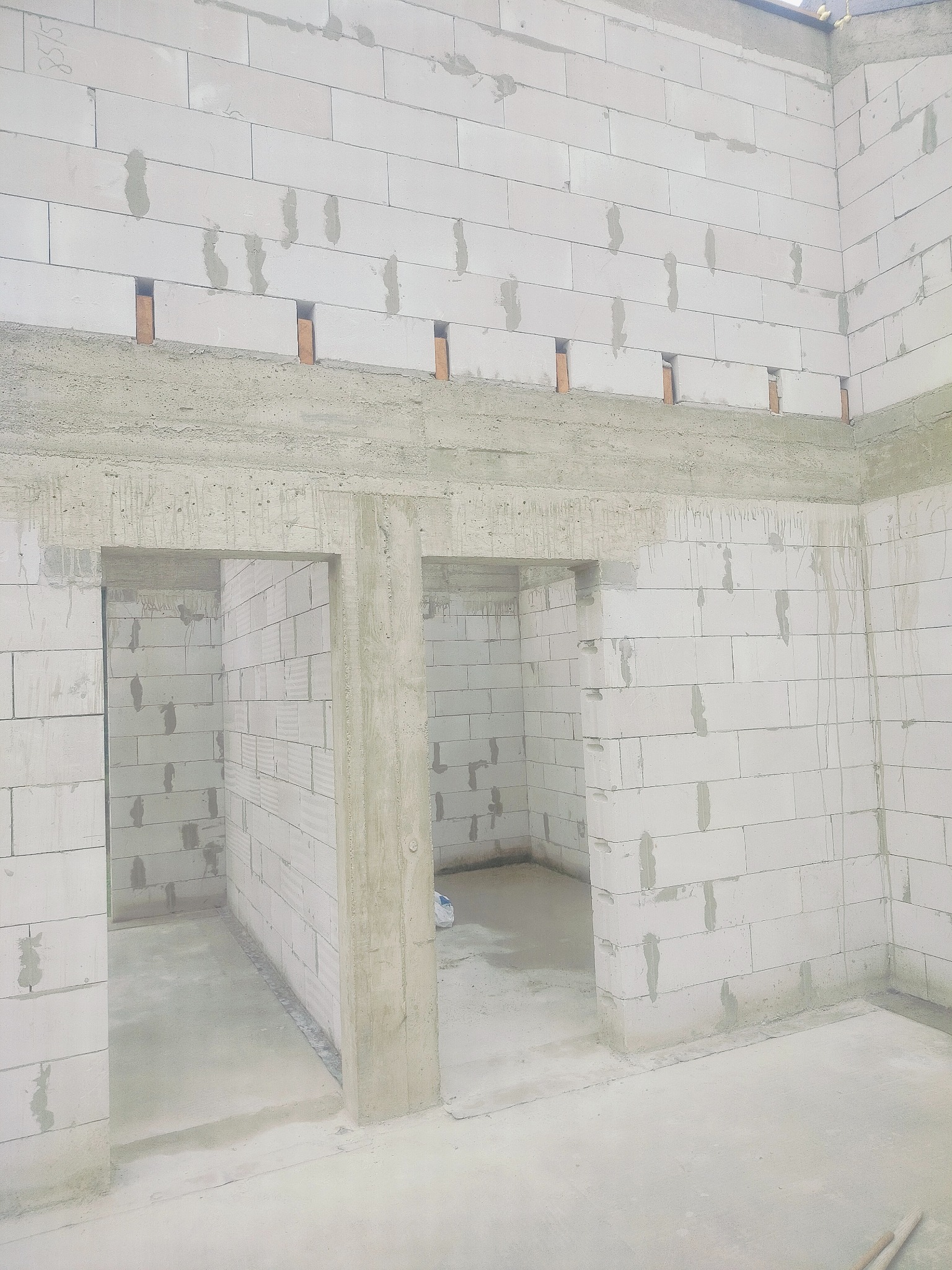 Wnętrze w trakcie budowy: ściany z białych bloczków, betonowe nadproża nad otworami drzwiowymi, surowy beton na podłodze. Widoczne niedoskonałości i ślady zaprawy.