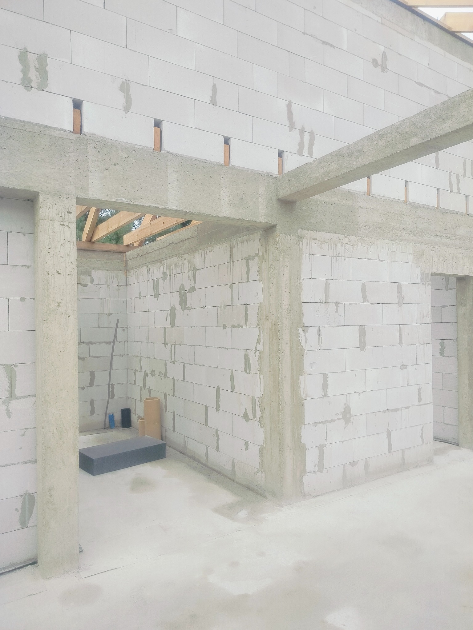 Wnętrze surowego domu w trakcie budowy: białe ściany z betonu komórkowego, betonowe nadproża i słupy, widoczne drewniane elementy więźby dachowej.