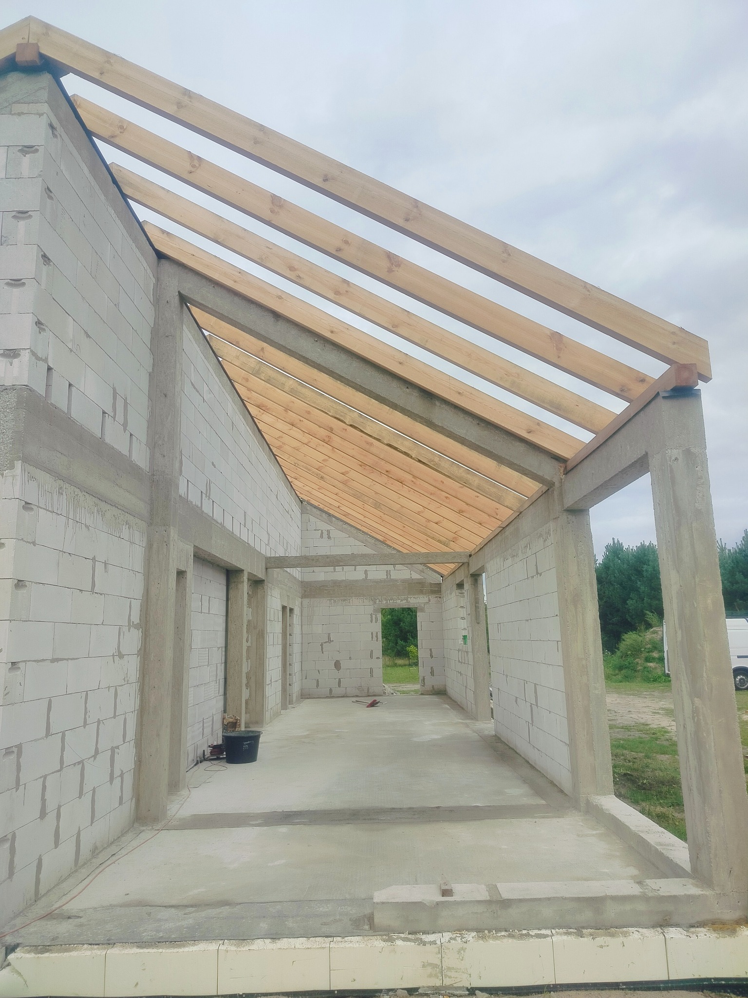 Surowy stan domu z betonu komórkowego z drewnianą konstrukcją dachu. Widoczne ściany, słupy i podłoga w szarym odcieniu. W tle zieleń drzew i fragment białego samochodu.