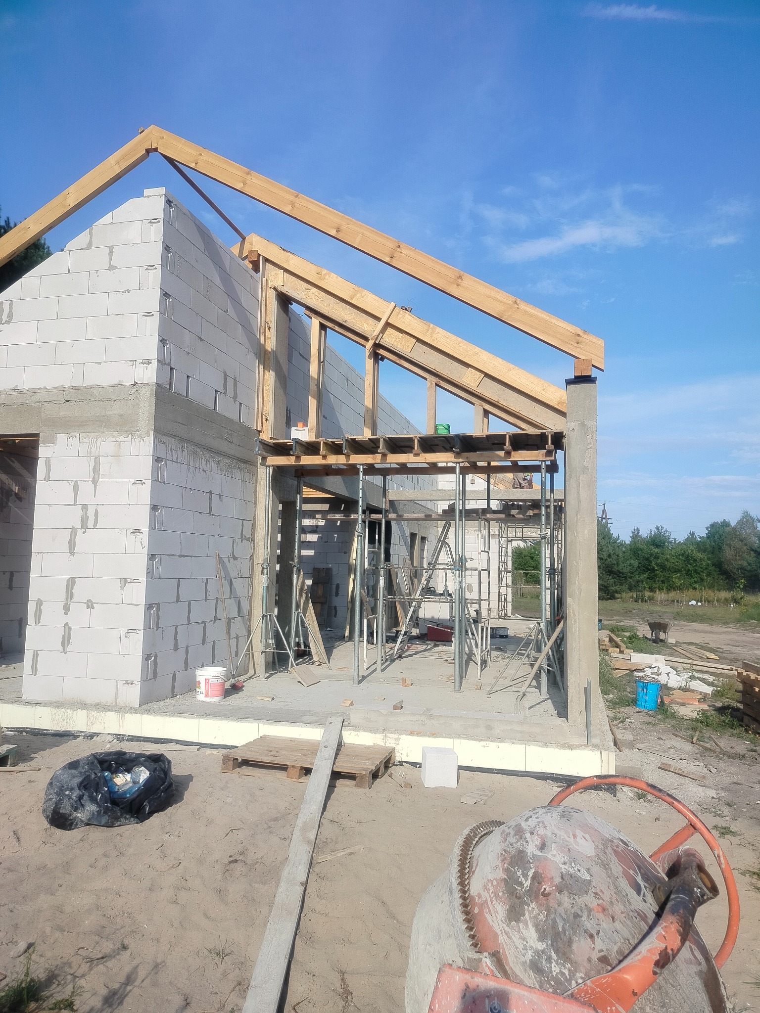 Surowy stan domu z białych bloczków, z widoczną konstrukcją dachu i rusztowaniami. Na pierwszym planie betoniarka i teren budowy.